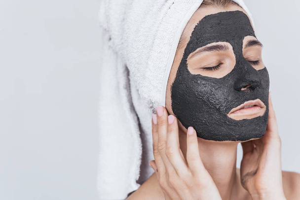 Closeup shot of young beautiful female applying black clay mask, doing beauty wellness treatment on her face skin. Horizontal shot of woman applying organic facial mask, with closed eyes on white - Foto, Imagen