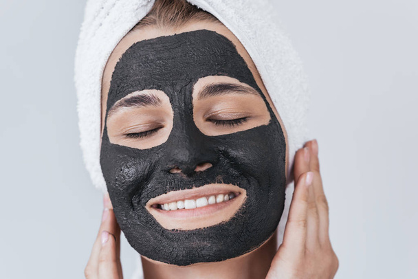 若い女性美しい黒い粘土マスク適用を笑っている顔の皮膚の美容健康治療を行うのクローズ アップ ショットをトリミングしました。目を閉じて、有機の顔のマスクを適用する女性の水平ショット. - 写真・画像
