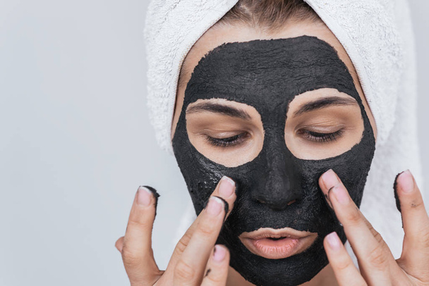 Closeup portrait of beautiful young woman applying clay black mask, doing beauty wellness treatment on her face skin. Horizontal shot of amazed female applying organic facial mask, looking down.  - Foto, afbeelding