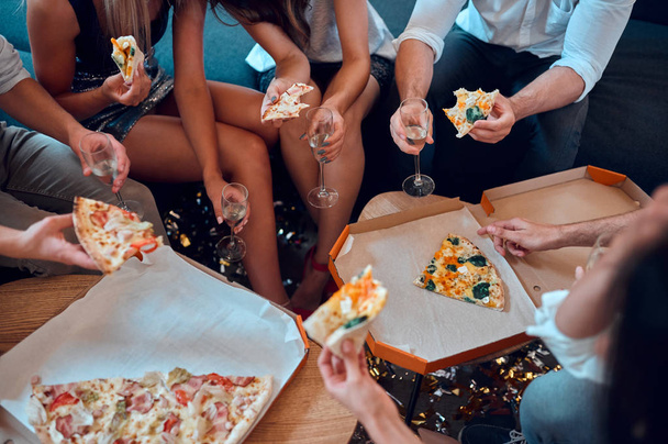 Let the party begin! Cropped image of group of young people having fun together. Sitting in big light room with champagne and pizza. Celebrating holiday in big company of close friends. - 写真・画像