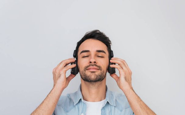 Portrait of handsome young Caucasian male with cheerful expression, closes eyes as feels enjoyment and relaxation, wearing blue shirt, holding headphones with both hands, listening music. Copy space - Foto, imagen