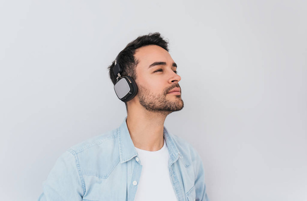 Portrait of serious handsome Caucasian male listens music or audio book, uses modern wireless headphones, poses against grey background with copy space for your information advertising or text.  - Fotografie, Obrázek