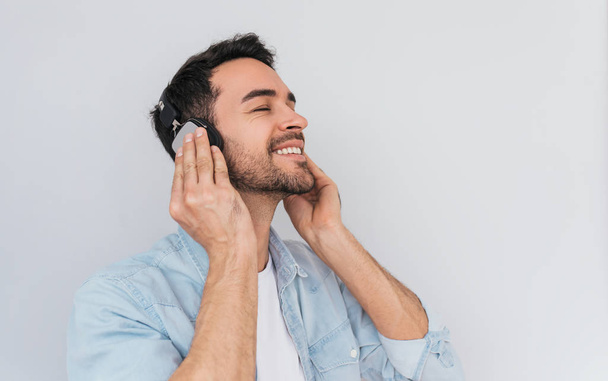 Horizontal portrait of happy young Caucasian male wearing in casual blue shirt, holding headphones with both hands, listening favorite music with closed eyes isolated on gray background. Copy space - Foto, immagini