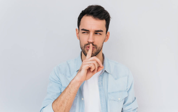 Portrait of serious fashionable male dressed trendy in blue shirt, keeps fore finger on lips, making shh gesture for secret or be quiet. Isolated shot of stylish man shows silence sign. People concept - Фото, зображення