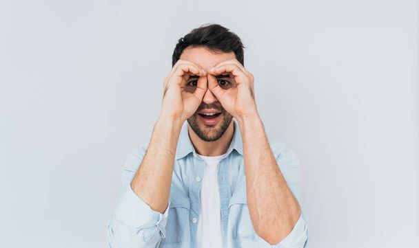 Handsome amazed male wearing blue shirt and white T-shirt holding his hands at his eyes as if looking through binoculars or glasses, surprised happily against studio light wall background. People - 写真・画像