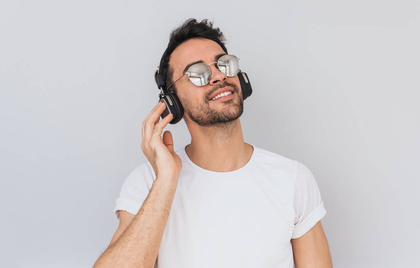 Studio horizontal portrait of happy young male wearing white tshirt and mirros glasses, holding headphones with hand, listening favorite music with closed eyes isolated on white background. Copy space - Photo, Image