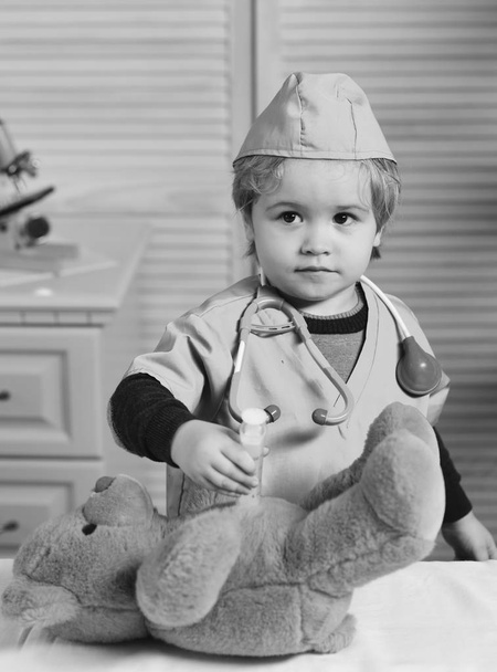 Kid in doctor coat makes injection to teddy bear. Boy in surgical uniform holds syringe on wooden background - Photo, image