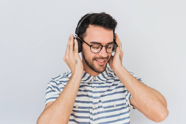 Horizontal shot of handsome happy young male wearing striped shirt and trendy glasses, with headphones on head, listening favorite music, isolated on white studio background.Copy space for advertising - Zdjęcie, obraz