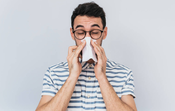 Horizontal portrait of unhealthy handsome man wearing striped shirt and glasses, blowing nose into tissue. Male have flu, virus or allergy against white background. Healthy medicine and people concept - Photo, image