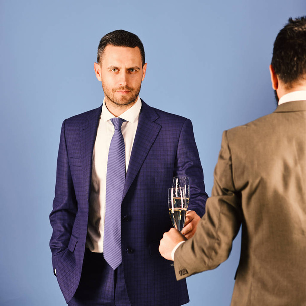 Men in smart suits on blue background. Businessman with smile holds sparkling wine clinking glasses - Zdjęcie, obraz