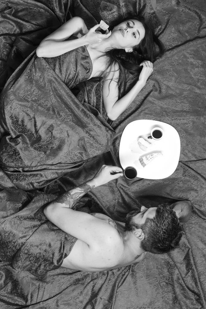 Man with beard has romantic breakfast with dreamy lady in bed, topview. Man and woman with half covered bodies drink coffee - Photo, Image