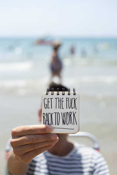 closeup of a young caucasina man in the beach showing a spiral notepad, in front of his face, with the text get the fuck back to work handwritten in it - Foto, afbeelding
