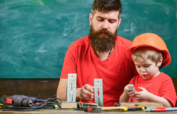 Boy, child in protective helmet makes by hand, repairing, does crafts with dad. Father, parent with beard and little son in classroom, chalkboard on background. Father as handyman concept - Foto, afbeelding