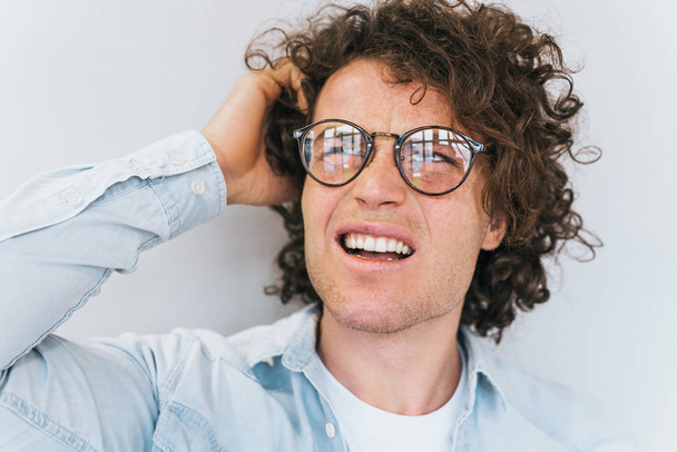 Cropped closeup portrait of handsome pensive male with curly hair and freckles, wears denim shirt and trendy round spectacles, looking up and thinking, holding his head on white wall. People concept. - Фото, изображение