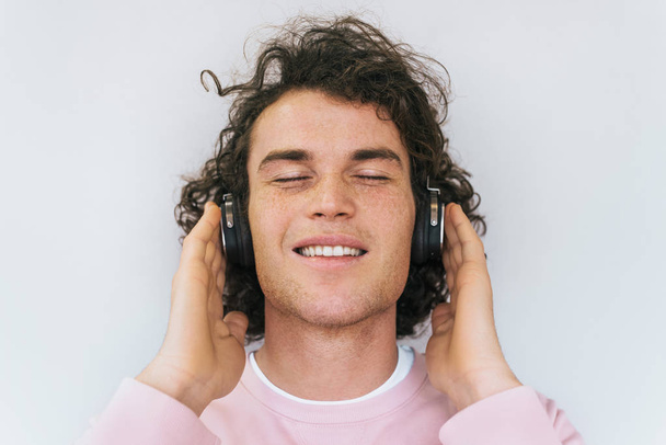 Closeup portrait of handsome smiling young male model with curly hair and freckles wearing pink clothes, listening music on headphones, isolated on white studio wall. Copy space for advertisement - Fotografie, Obrázek