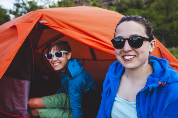 Kaksi tyttöä istuu teltassa. Tyttöystävät kommunikoivat luonnossa. Makuupusseissa olevat naiset juovat kahvia. Ystäviä leirintäalueella. Hymyilevät brunetit puhuvat
. - Valokuva, kuva