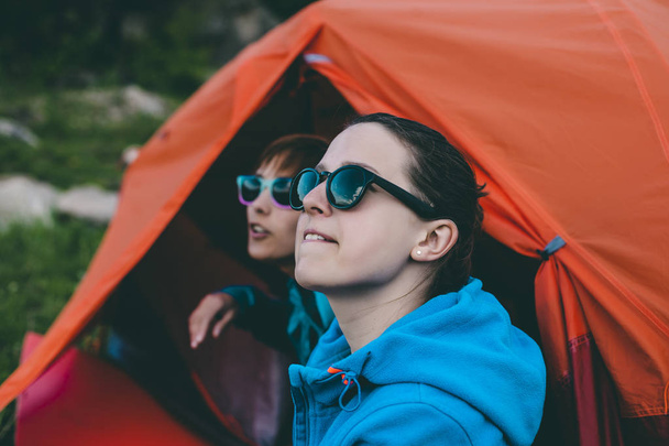 Kaksi tyttöä istuu lähellä telttaa. Tyttöystävät kommunikoivat luonnossa. Ystävien kesken. Lepää kaupungin ulkopuolella. Naiset ihailevat auringonlaskua. Telttailu lähellä jokea
. - Valokuva, kuva