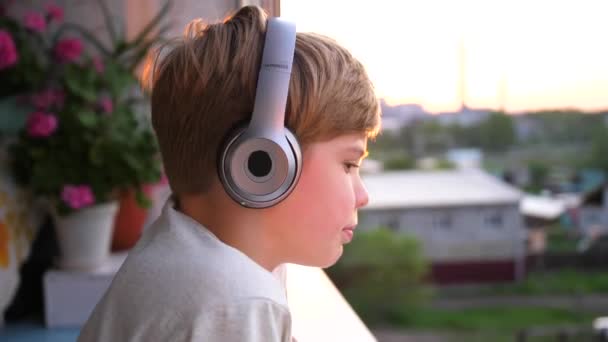 Egy fiatalember áll az ablak mellett, és felteszi a fejhallgatót, hogy zenét hallgasson. Homályos háttér naplementével, tini élvezi a zenét a fejhallgató. - Felvétel, videó