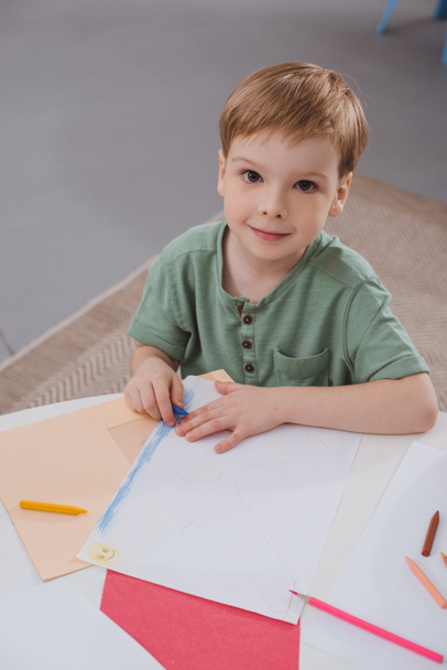 high angle view of preschooler boy sitting at table with paper and colorful pencils for drawing in classroom - Photo, image