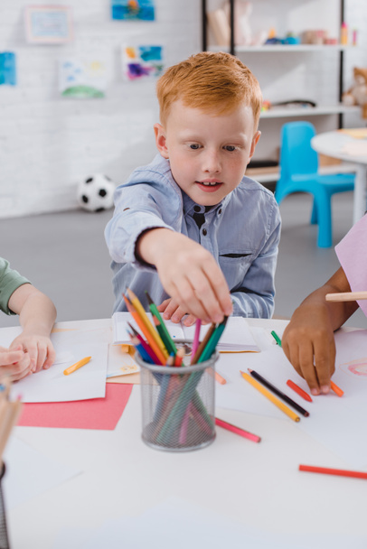 szelektív összpontosít, vörös hajú fiú vesz ceruza asztalnál multikulturális osztálytársai osztályteremben - Fotó, kép