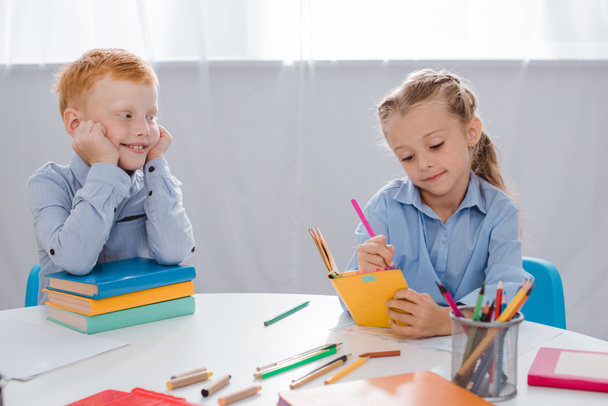 Porträt eines lächelnden Jungen mit rotem Haar, der einen Mitschüler ansieht, der im Klassenzimmer in ein Notizbuch schreibt  - Foto, Bild