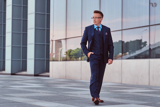 Уверенный бизнесмен, одетый в элегантный костюм, смотрит в сторону и держит руку в кармане во время прогулки по улице на фоне городского пейзажа
. - Фото, изображение