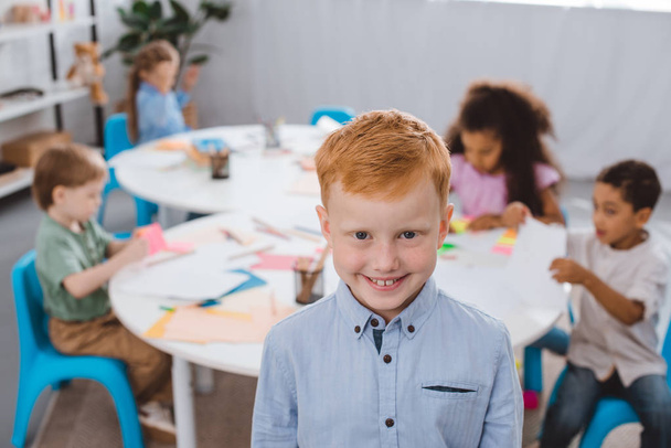 selective focus of happy red hair boy looking at camera with multiracial classmates behind in classroom - Fotografie, Obrázek