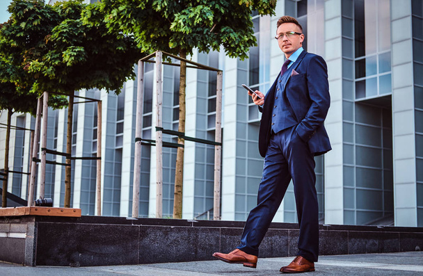Portrait of a confident stylish businessman dressed in an elegant suit holds a smartphone and looking away while standing outdoors against a skyscraper background. - Фото, зображення