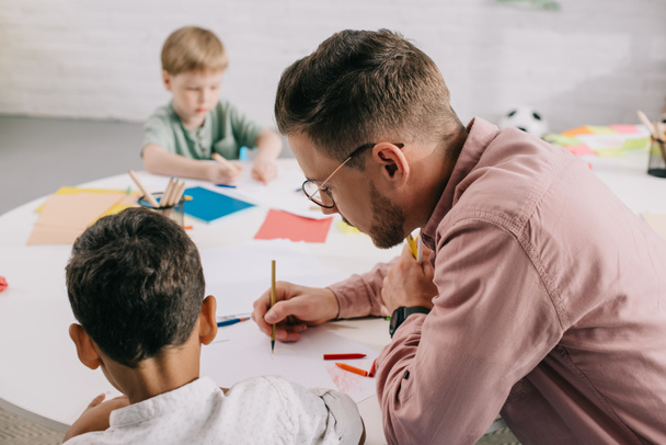 selective focus of teacher and multiracial boys drawing pictures with colorful pencils at table in classroom - Foto, Imagen