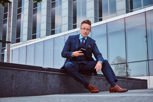Portrait d'un homme d'affaires élégant et confiant vêtu d'un costume élégant tient un smartphone et regardant la caméra assis à l'extérieur sur un fond de gratte-ciel
. - Photo, image
