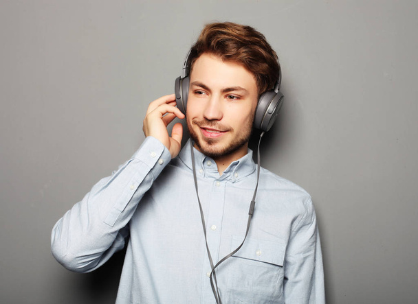 life style, happiness, emotional and people concept: Enjoying his favorite music. Happy young stylish man adjusting his headphones ad smiling while standing against grey background - Foto, Bild