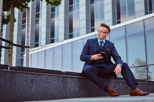 Retrato de un hombre de negocios con estilo y confianza vestido con un traje elegante sostiene un teléfono inteligente y mirando hacia otro lado mientras está sentado al aire libre contra un fondo de rascacielos
. - Foto, imagen