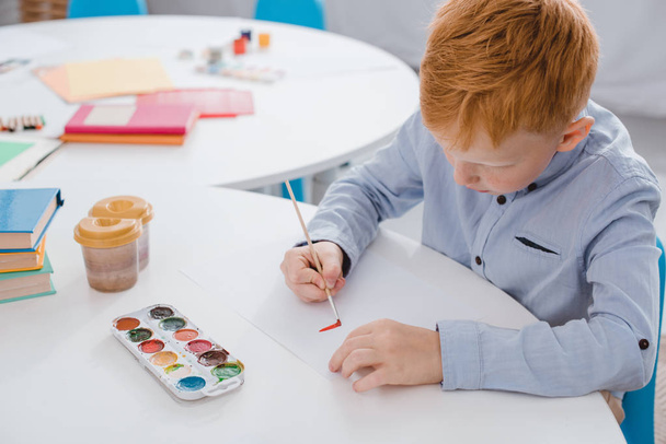 focused preschooler red hair boy drawing picture at table in classroom - Photo, Image
