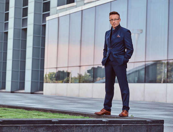 Full body portrait of a confident man in an elegant suit standing with hands in pockets against a skyscraper background. - Fotografie, Obrázek