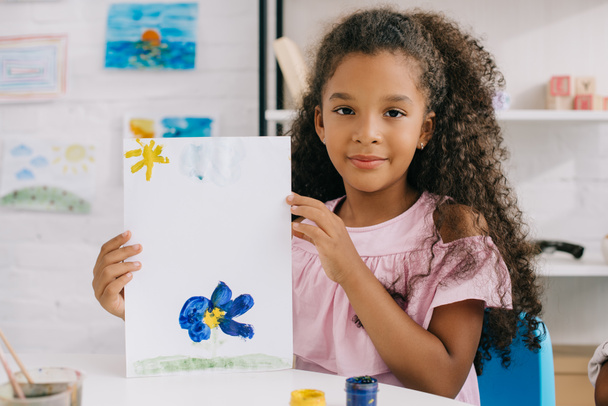 портрет афроамериканської дитини, що показує барвисту картину в руках, сидячи за столом в кімнаті
 - Фото, зображення
