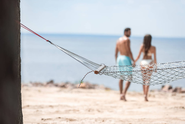 selective focus of couple holding hands and walking on beach with hammock on foreground - Photo, Image