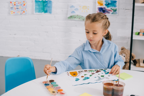 portrait of adorable focused child drawing colorful picture with paints and brush at table - Foto, Imagen