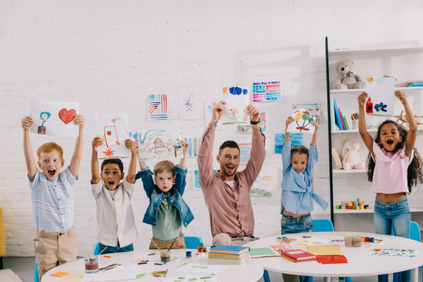 πορτρέτο του χαμογελώντας δάσκαλος και πολυεθνική παιδιά δείχνουν εικόνες στα χέρια στην τάξη - Φωτογραφία, εικόνα