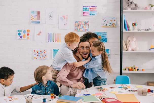 niños interracial abrazando feliz profesor en la mesa en el aula
 - Foto, Imagen