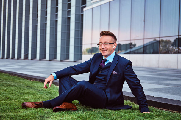 Happy businessman dressed in elegant suit sitting on a green lawn against cityscape background. - Photo, image
