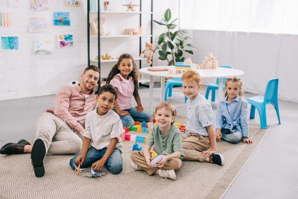 χαμογελώντας δάσκαλος και πολυπολιτισμική preschoolers κάθεται στο πάτωμα με τα πολύχρωμα τουβλάκια στην τάξη - Φωτογραφία, εικόνα