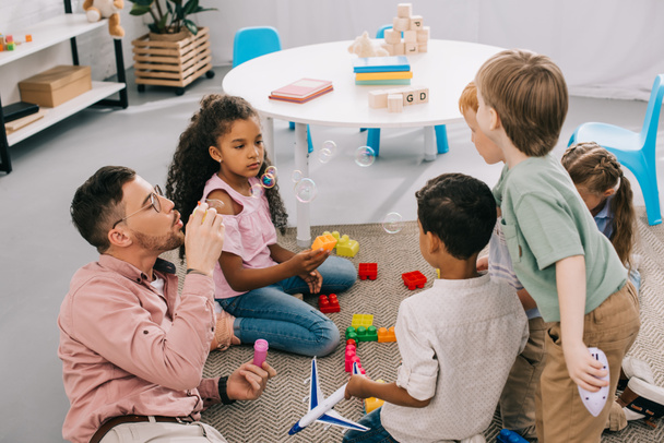 δάσκαλος με σαπουνόφουσκες και πολυπολιτισμική preschoolers κάθεται στο πάτωμα με τα πολύχρωμα τουβλάκια στην τάξη - Φωτογραφία, εικόνα