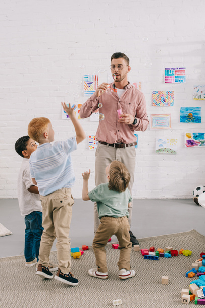 teacher blowing soap bubbles to multiracial boys in classroom - Photo, Image