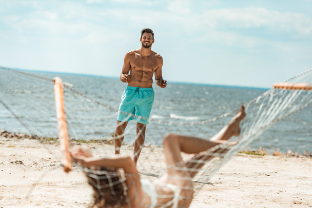 komea paidaton mies tuo kookoscocktaileja tyttöystävälleen riippumatossa lähellä merta
 - Valokuva, kuva