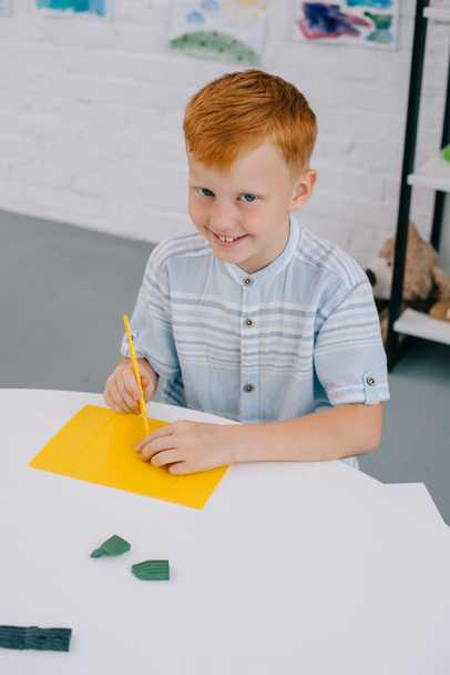 high angle view of smiling red hair boy sitting at table with colorful plasticine for sculpturing in room - Photo, image