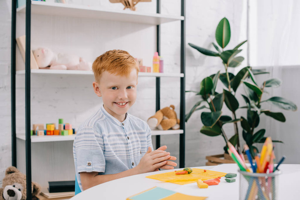 portrait of smiling red hair boy sitting at table with colorful plasticine for sculpturing in room - Photo, image