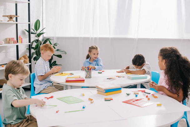 милые мультиэтнические дошкольники, скульпторы с пластилином за столами в классе
 - Фото, изображение