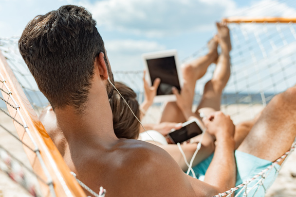 νεαρό ζευγάρι ακούει μουσική με ακουστικά και gadgets ενώ χαλαρώνει στην αιώρα στην παραλία - Φωτογραφία, εικόνα