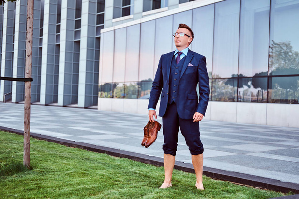 Confident businessman dressed in elegant suit holds his shoes enjoying while standing barefoot on a green lawn against cityscape background.  - 写真・画像