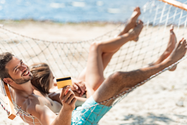 海の近くに金色のクレジット カードを保持していると、ビーチでハンモックに横になっている若いカップル - 写真・画像
